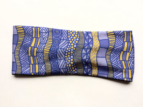 Stretchy headband featuring a soft bamboo liner and performance fabric outer shell. Light purple and gold bands of geometric patterns circle the headband
